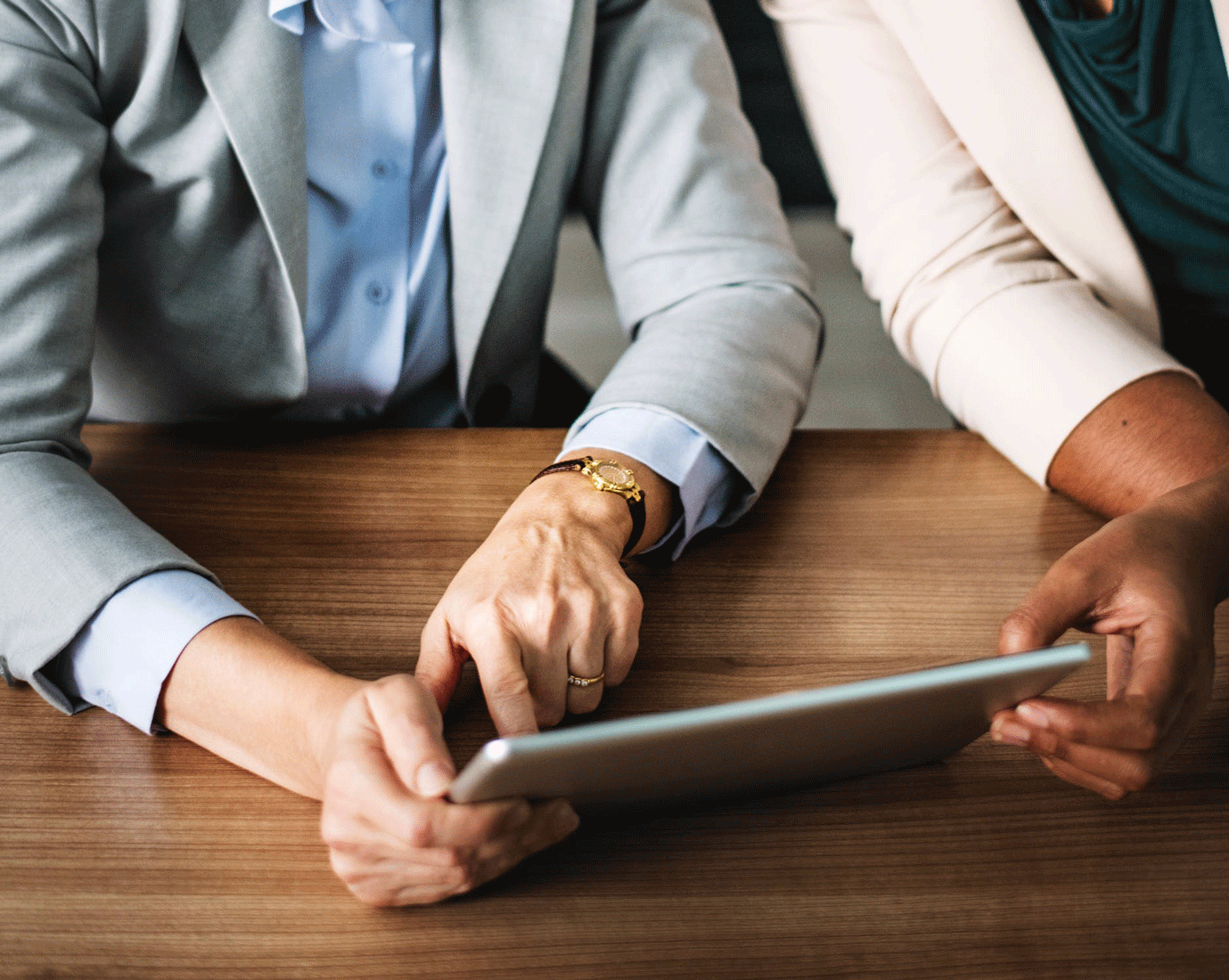 Two people in business attire sit at a wooden table; one points at a tablet while the other holds it, both looking at the screen.
