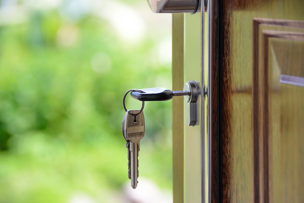 Close-up of a keychain with two keys in the lock of a wooden door, with a blurred green outdoor background.