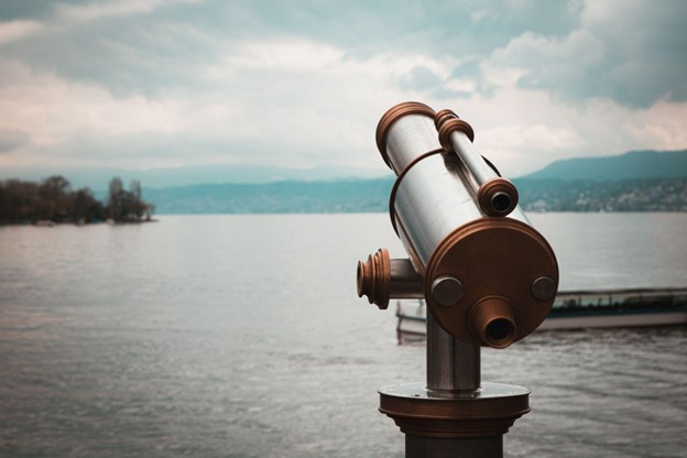 A metal public viewing telescope faces a large body of water, with distant mountains, cloudy skies, and a boat at right.