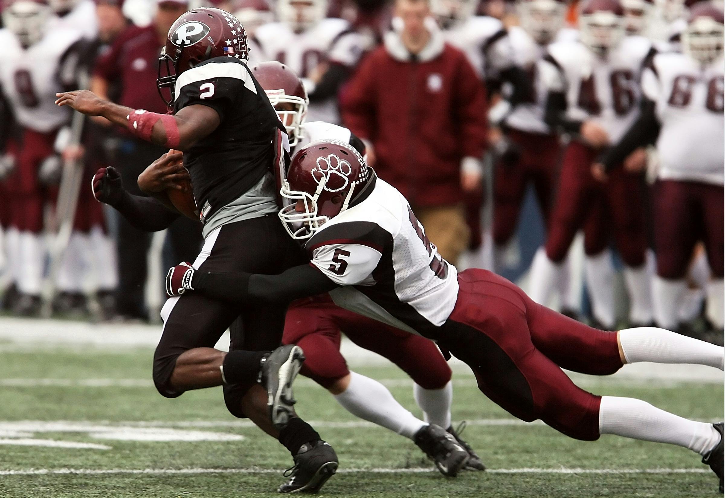 A football player in a black uniform runs with the ball as a maroon and white player tackles him from behind on a grassy field.