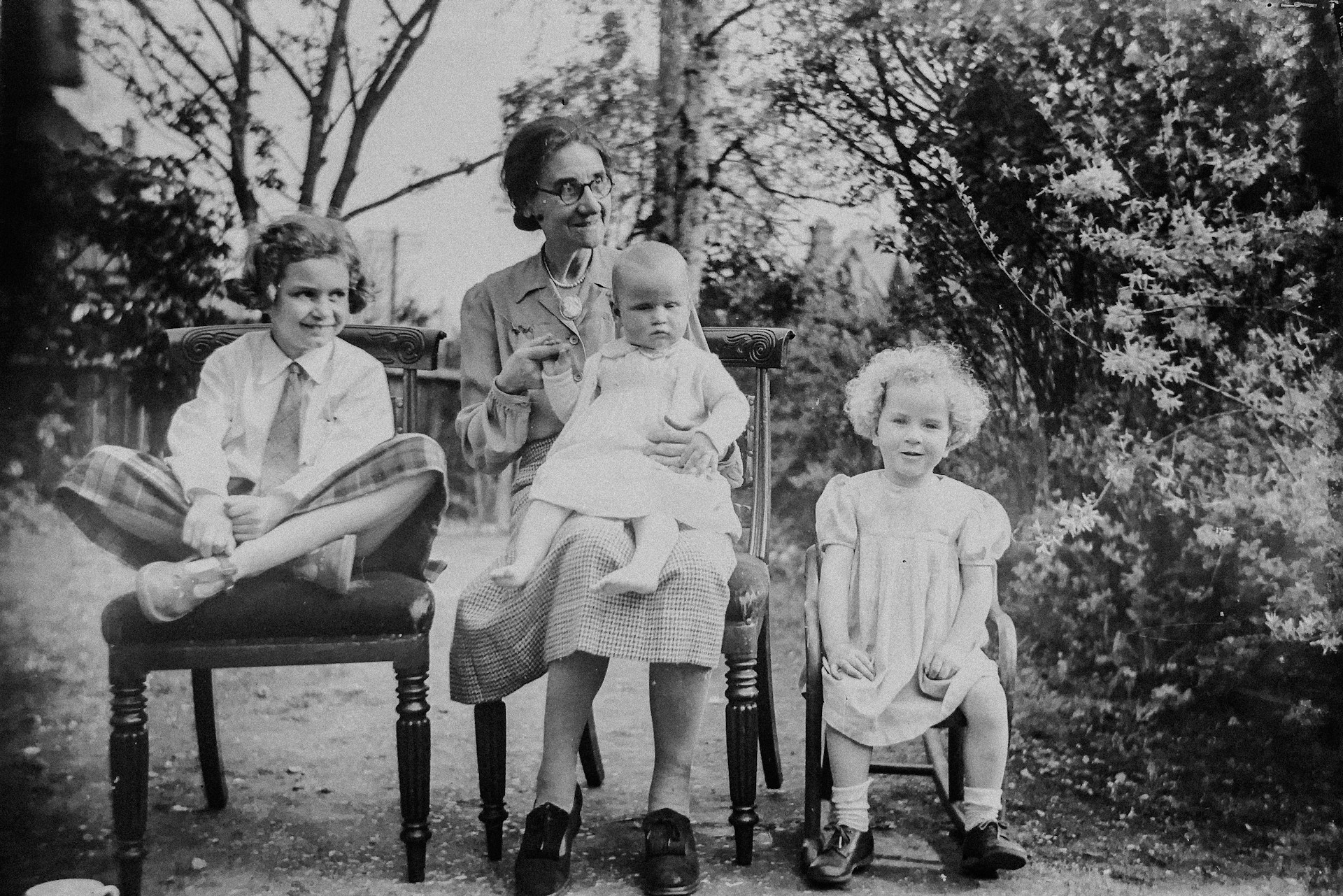 A black-and-white photo of a woman sitting outdoors with three children, one on her lap and two beside her, amid trees and foliage.