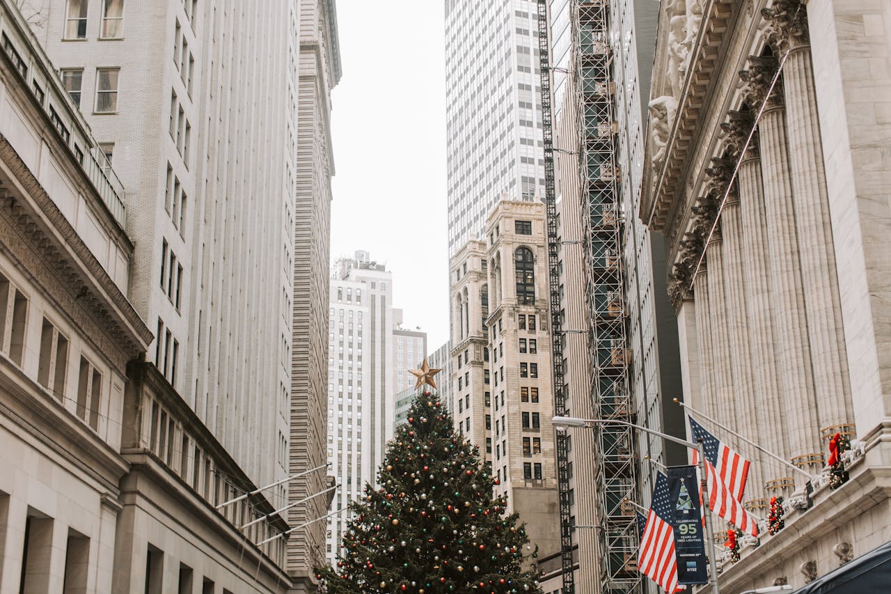 A large decorated Christmas tree stands in a city street, flanked by tall buildings, American flags, and festive wreaths.