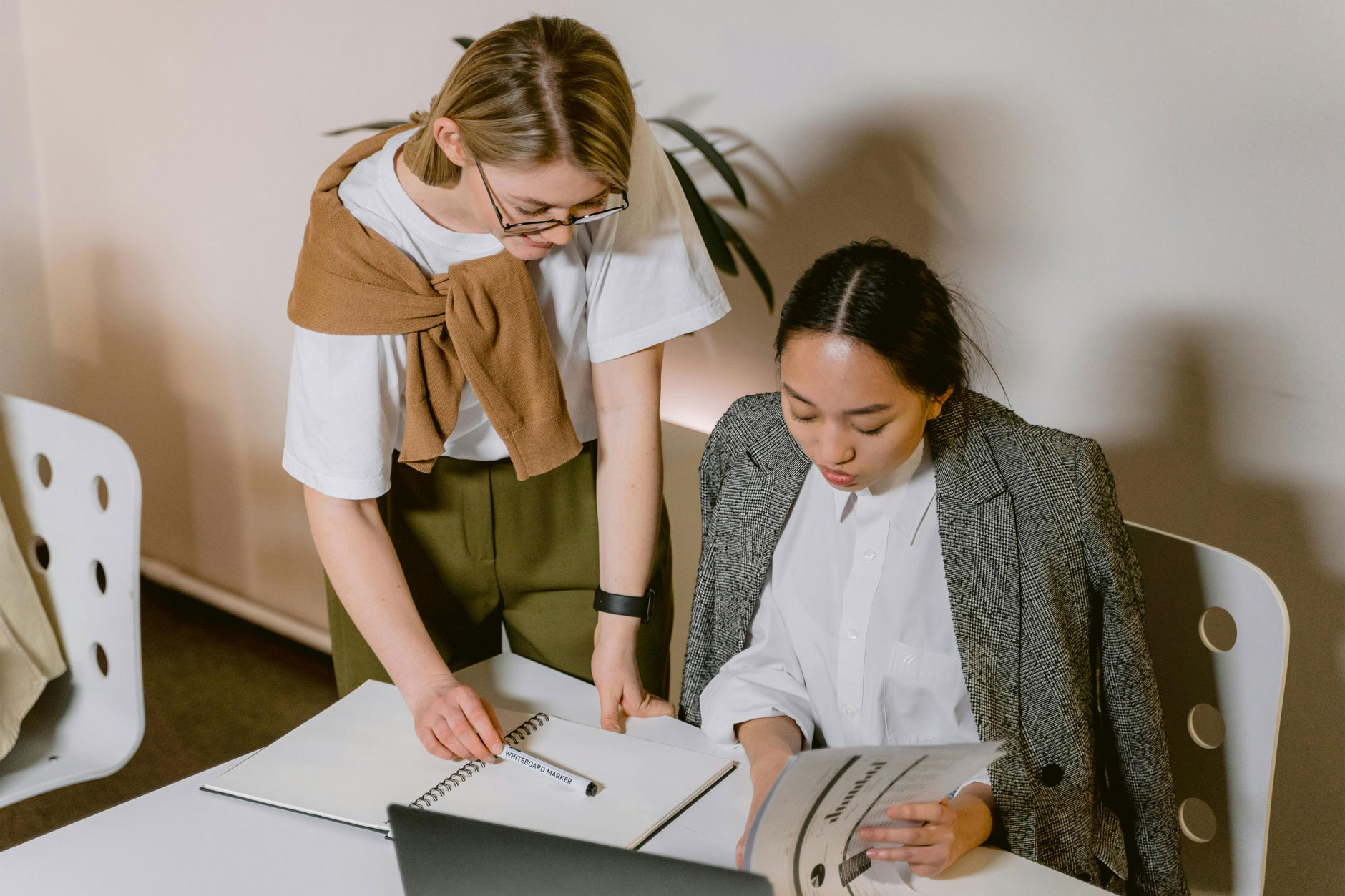 Two women at a table collaborate on estate research; one stands pointing at a notebook, the other sits reading a document.