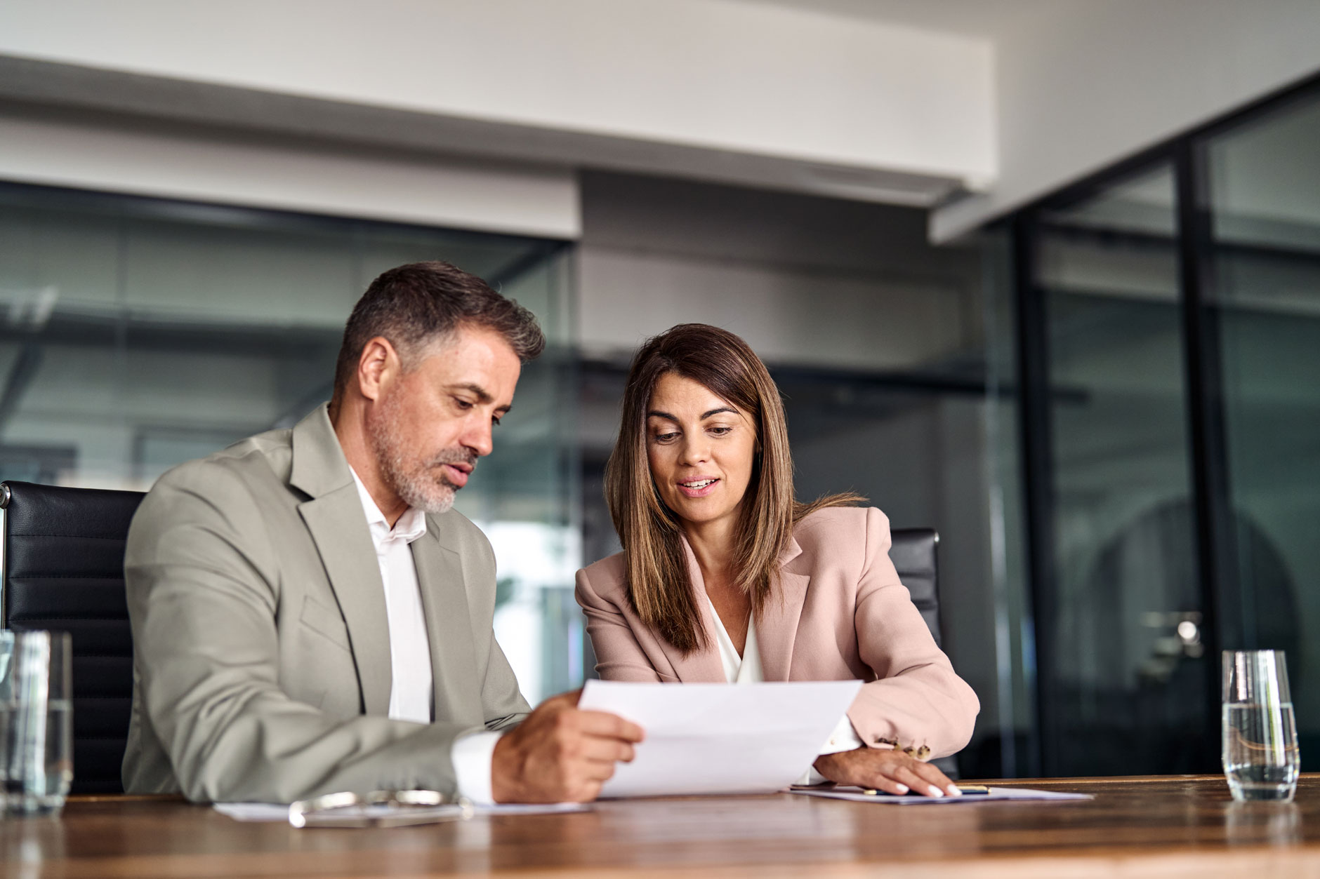 A man and a woman in business attire review documents together at a conference table in a bright, modern office.