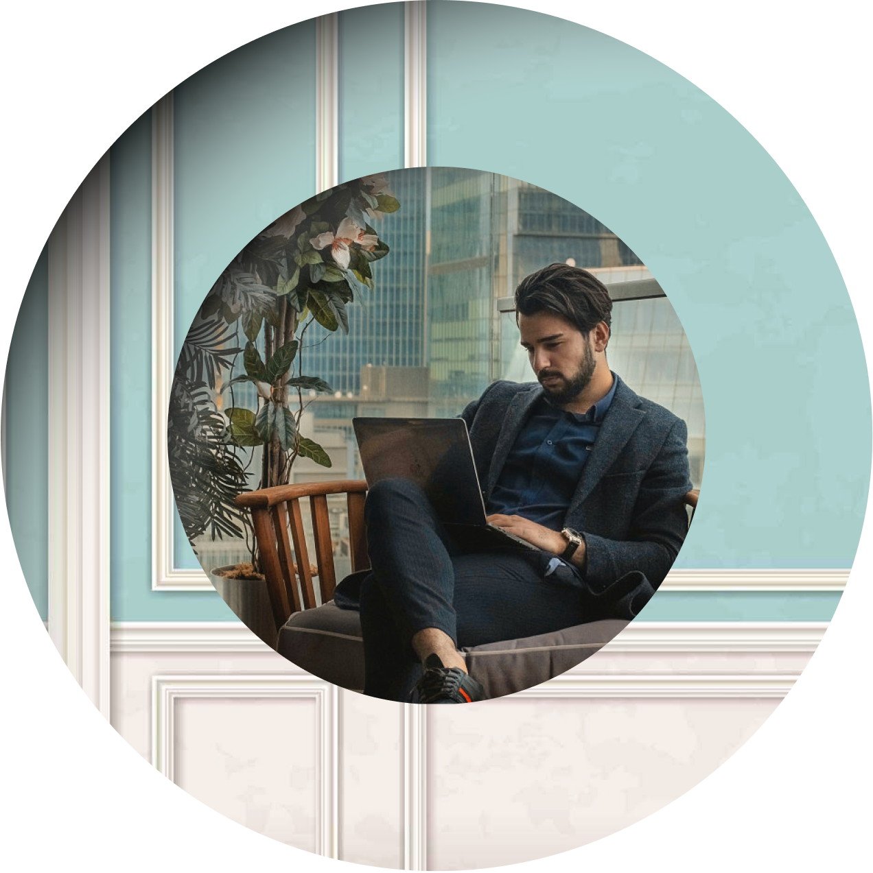 A man in a dark suit sits indoors on a chair, working on his laptop. City buildings and a large plant are visible nearby.