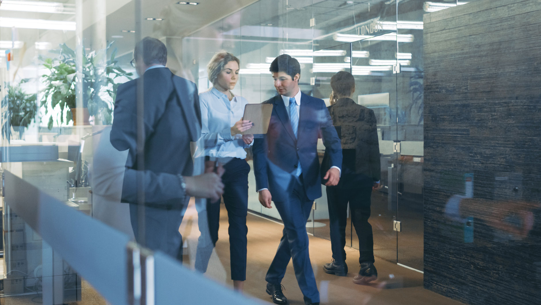 Two business professionals walk down a bright, glass-walled office hallway, reviewing a document on a clipboard. Others work nearby.