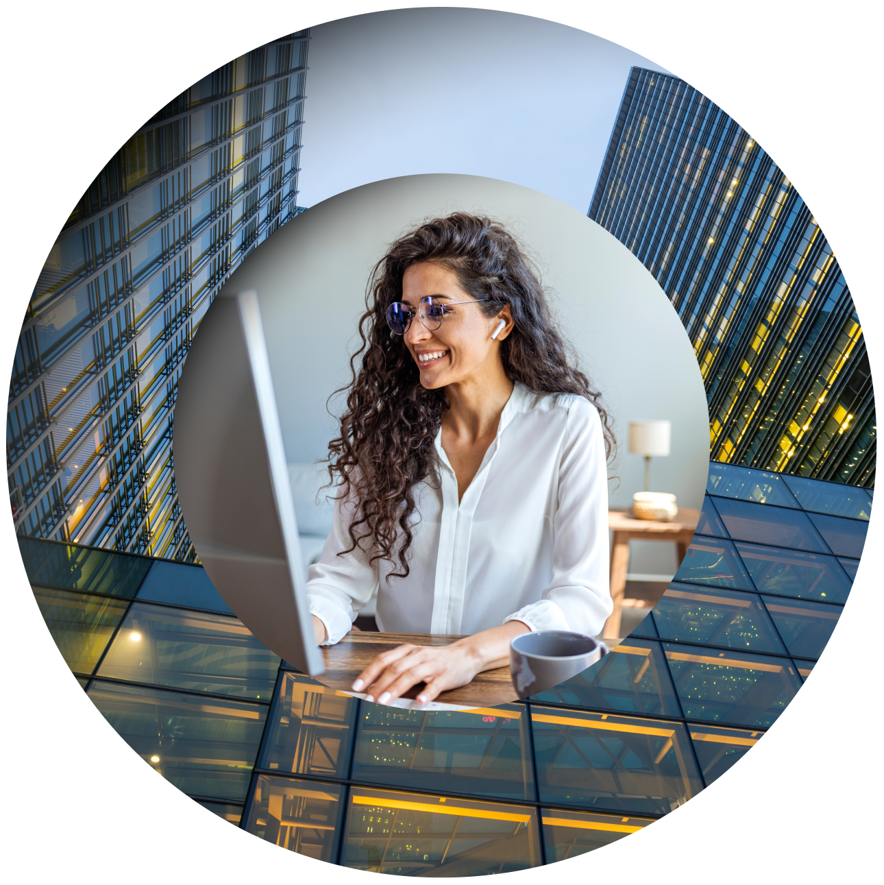 A woman with curly hair and glasses smiles while working on a laptop at a desk, with glass office buildings behind her.