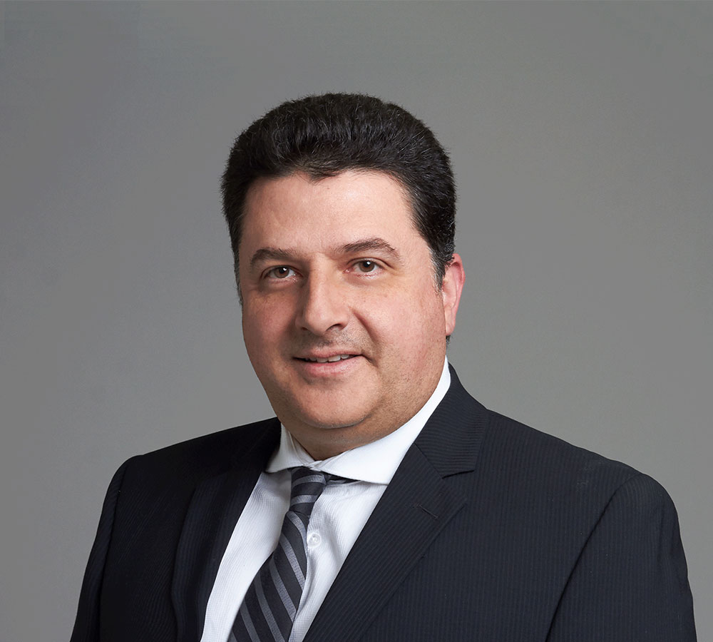 A man with short dark hair in a black suit, white shirt, and striped tie stands against a plain gray background.