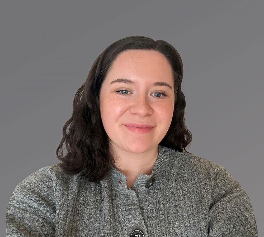 A woman with shoulder-length dark brown hair and fair skin smiles, wearing a textured gray cardigan against a plain gray background.