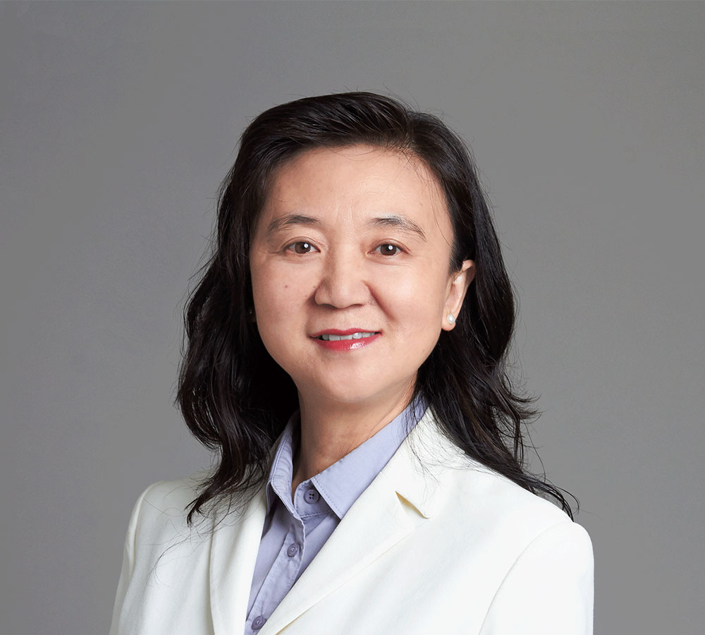 A woman with long, wavy black hair in a white blazer over a light blue shirt smiles softly against a plain gray background.