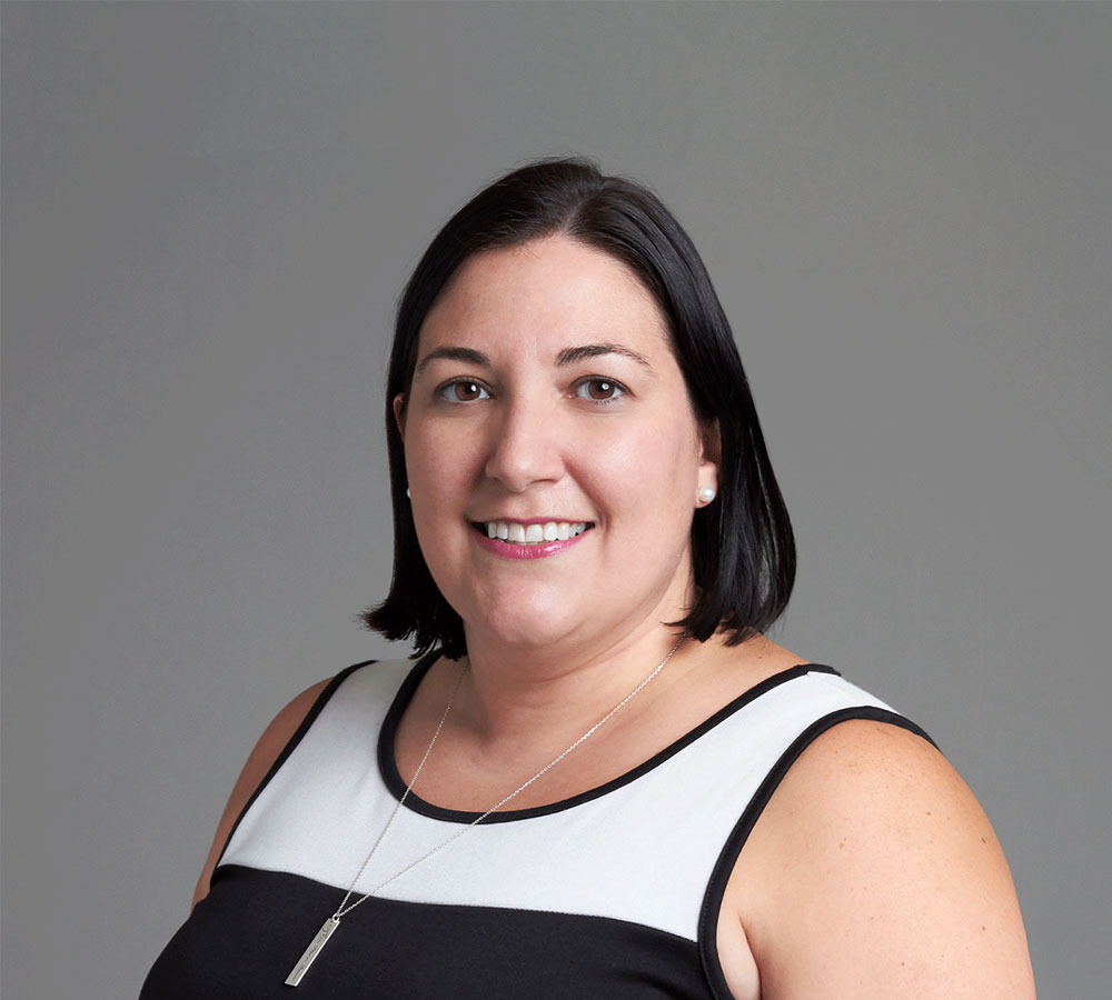 A woman with straight, dark shoulder-length hair in a sleeveless black and white top and necklace, smiling at the camera.