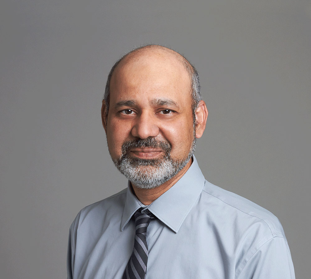 A middle-aged man with a beard and mustache, in a light blue dress shirt and striped tie, smiles slightly against a gray background.
