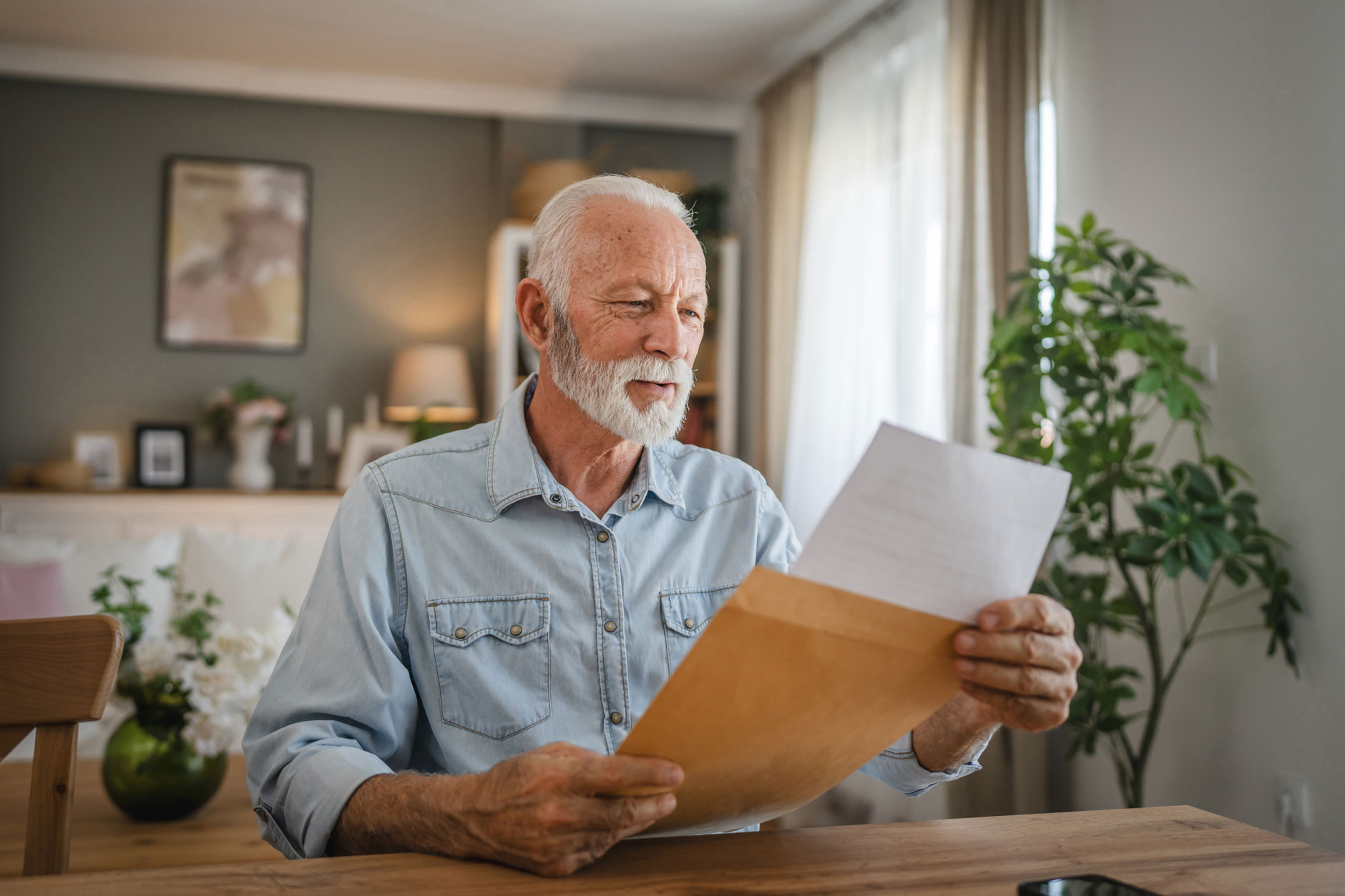 An older man with white hair and a beard smiles as he reads a letter at a wooden table in a sunlit, cozy living room.