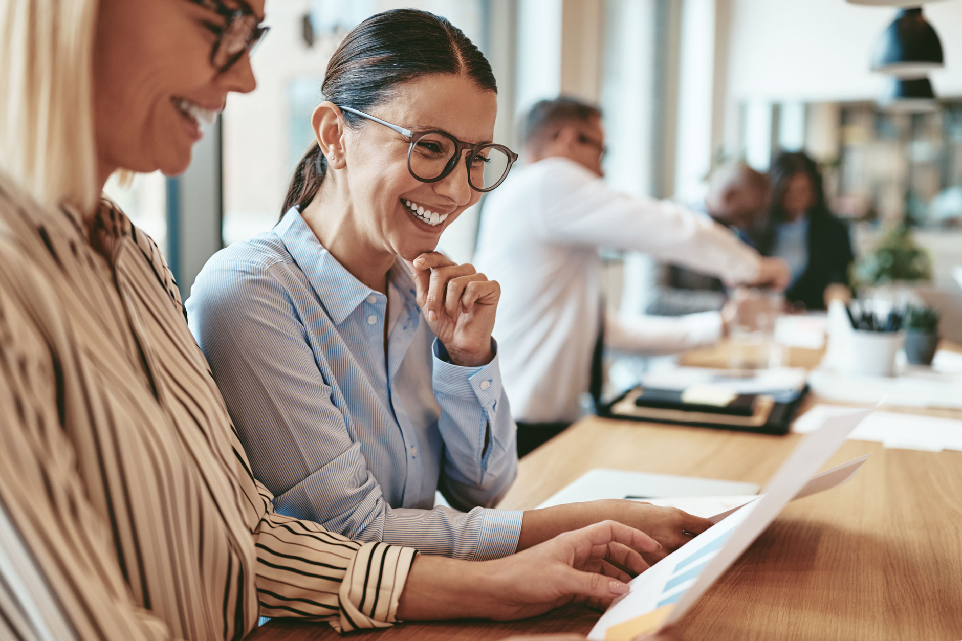 Two women in business attire smile as they review documents at a desk in a bright office, with colleagues working behind them.