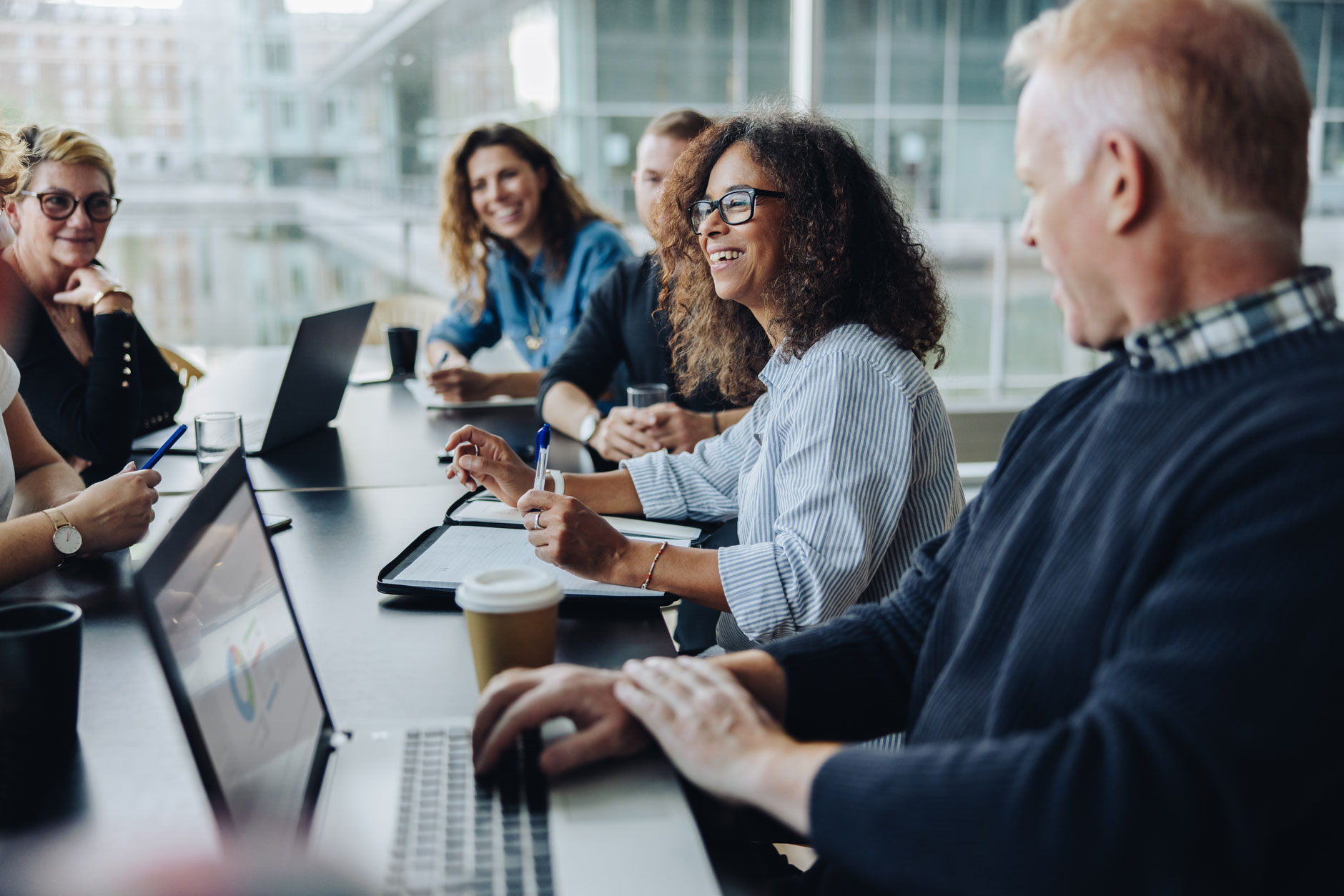 Five people sit around a table with laptops, notebooks, and coffee cups, talking in a modern office with large windows.