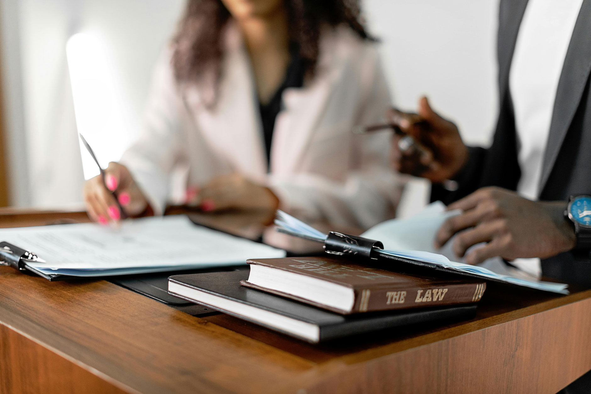 Two people sit at a wooden desk reviewing documents, with a book titled "THE LAW" and a clipboard visible in the foreground.
