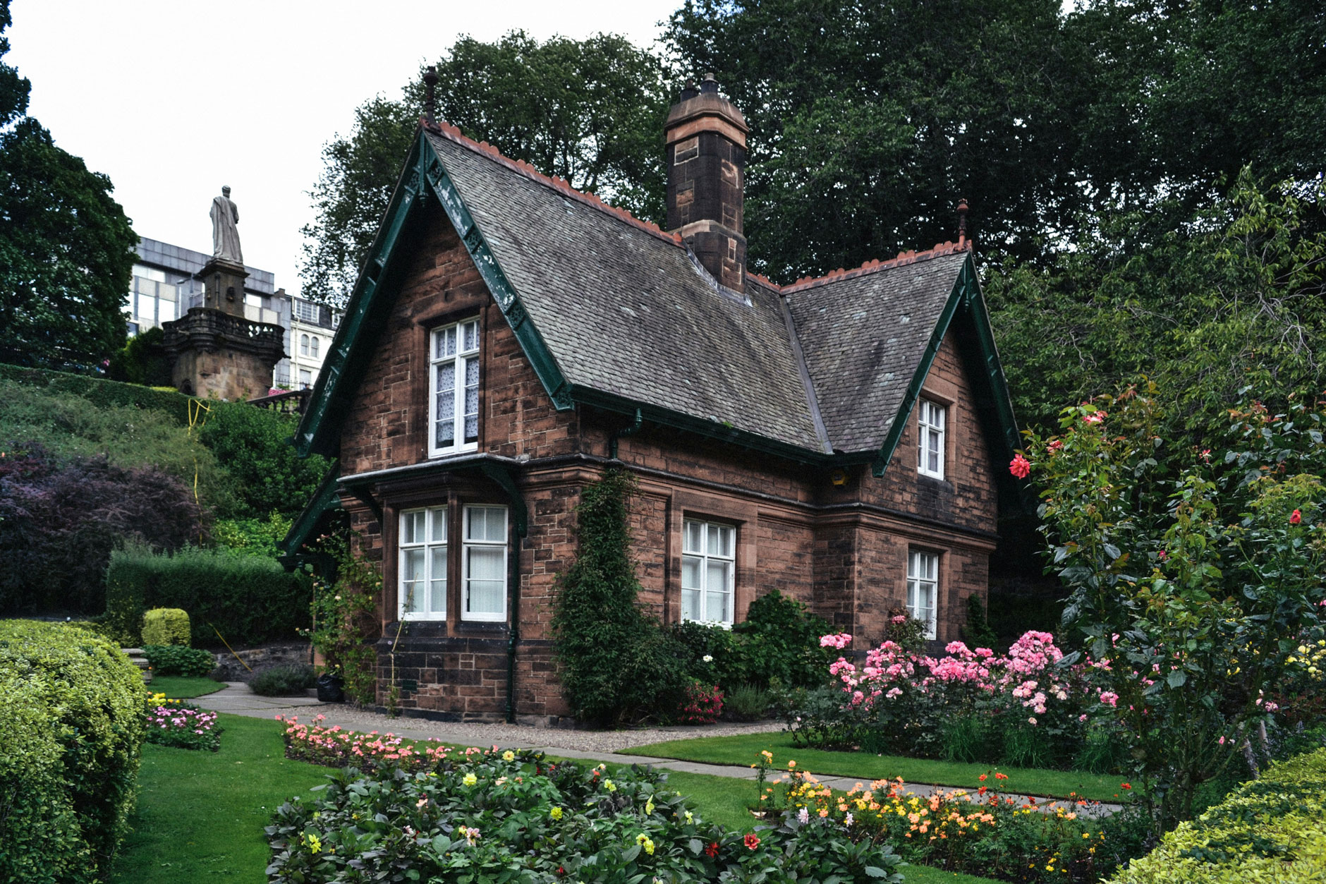 A brick cottage with a steep roof stands in a vibrant garden of flowers and trees; a stone statue is visible on a hill behind.