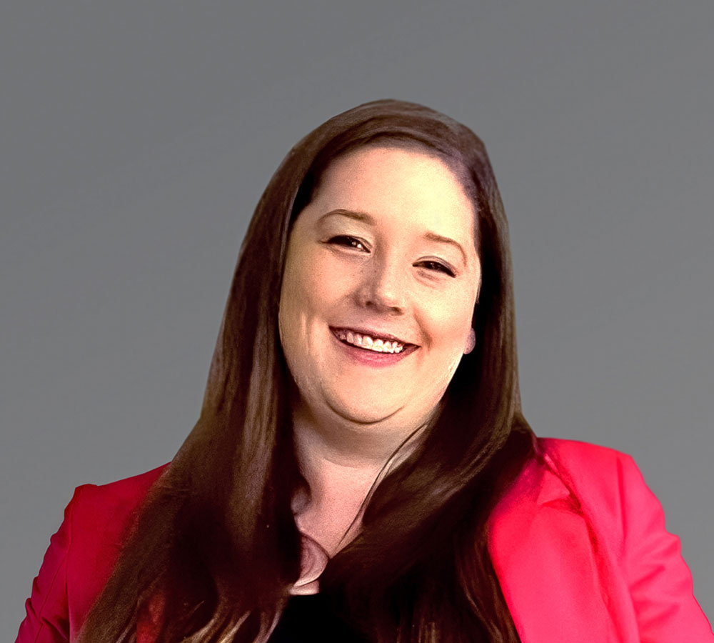 A woman with long brown hair smiles, wearing a bright pink blazer over a black top, in front of a plain gray background.