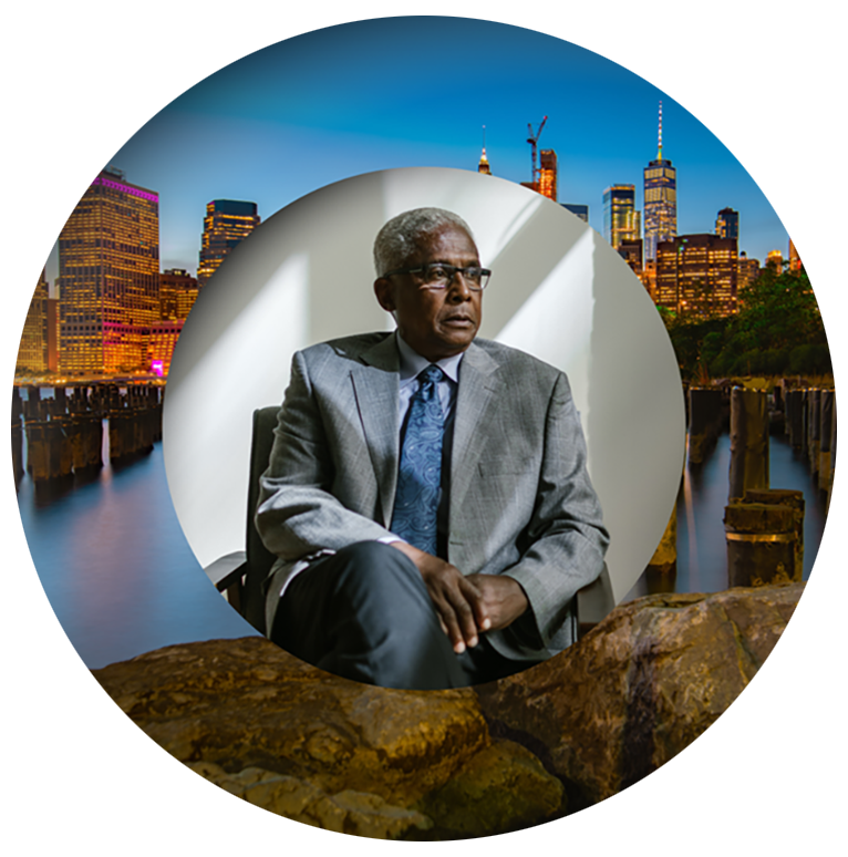 An older man in a gray suit and tie sits in a chair, overlaid on a circular dusk city skyline with water and wooden posts.