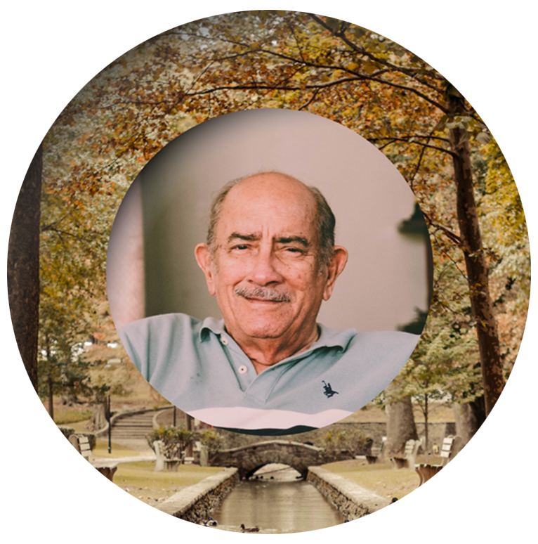 An elderly man with a mustache, in a light blue polo, smiles in front of an autumn park with a stone bridge and colorful trees.