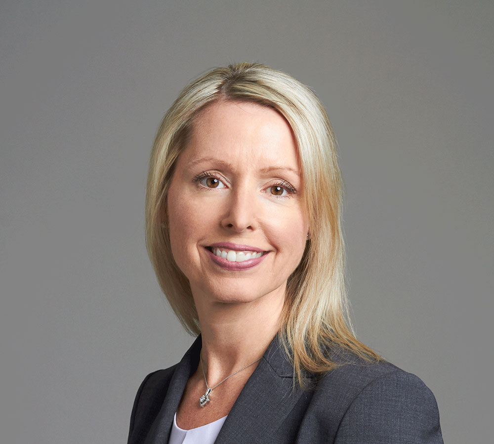A woman with straight blonde hair, in a grey blazer, white top, and silver necklace, smiles at the camera against a plain grey background.