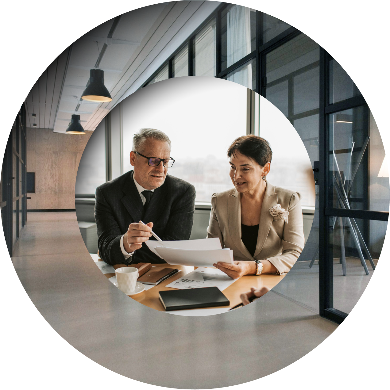 A man and woman sit at a table in a modern office with laptops, documents, and coffee cups, framed by a circular overlay.