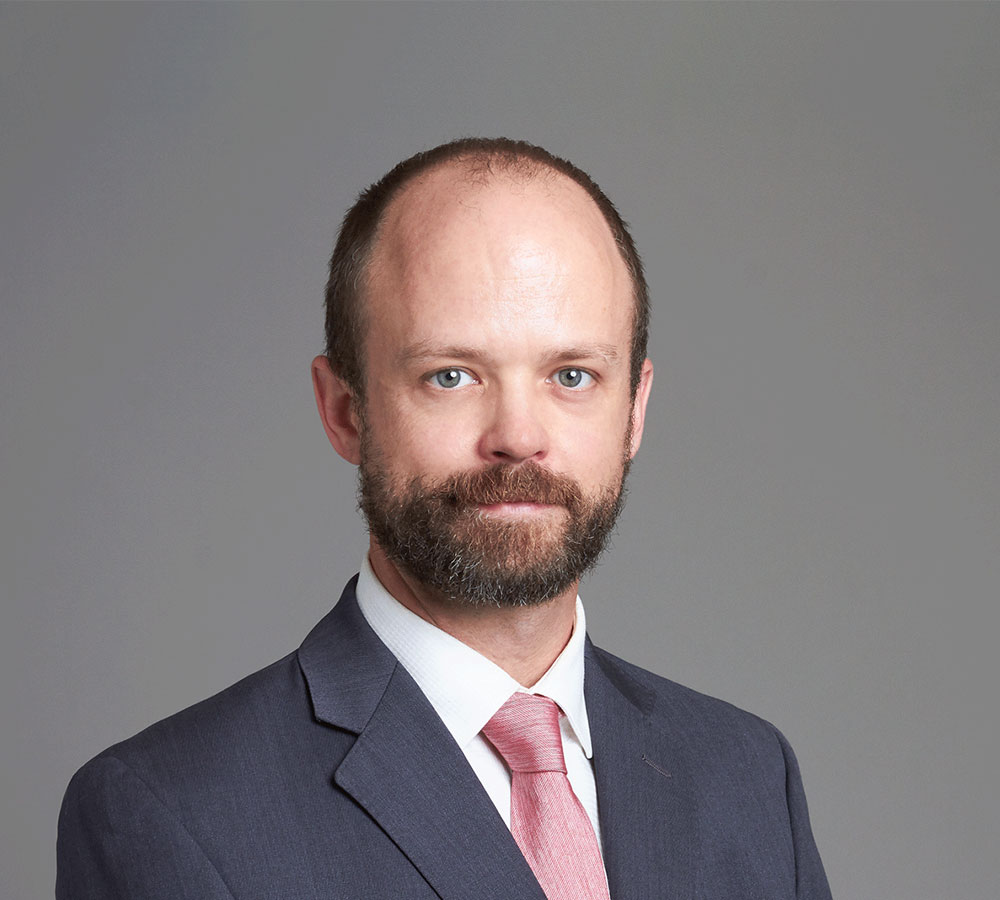 A bearded man in a dark suit, white shirt, and pink tie poses formally against a plain gray background.
