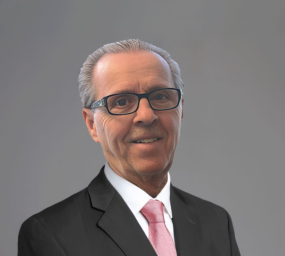 A middle-aged man with short gray hair, glasses, and a black suit with a light pink tie smiles before a plain gray background.