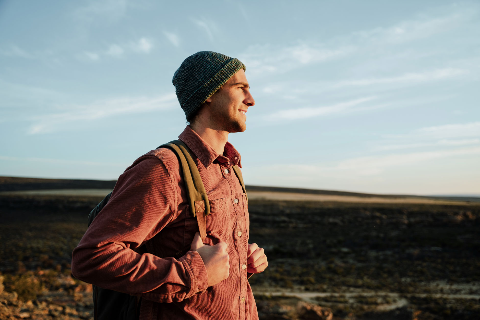 A person in a beanie and backpack stands in open fields at sunrise or sunset, smiling slightly while looking into the distance.