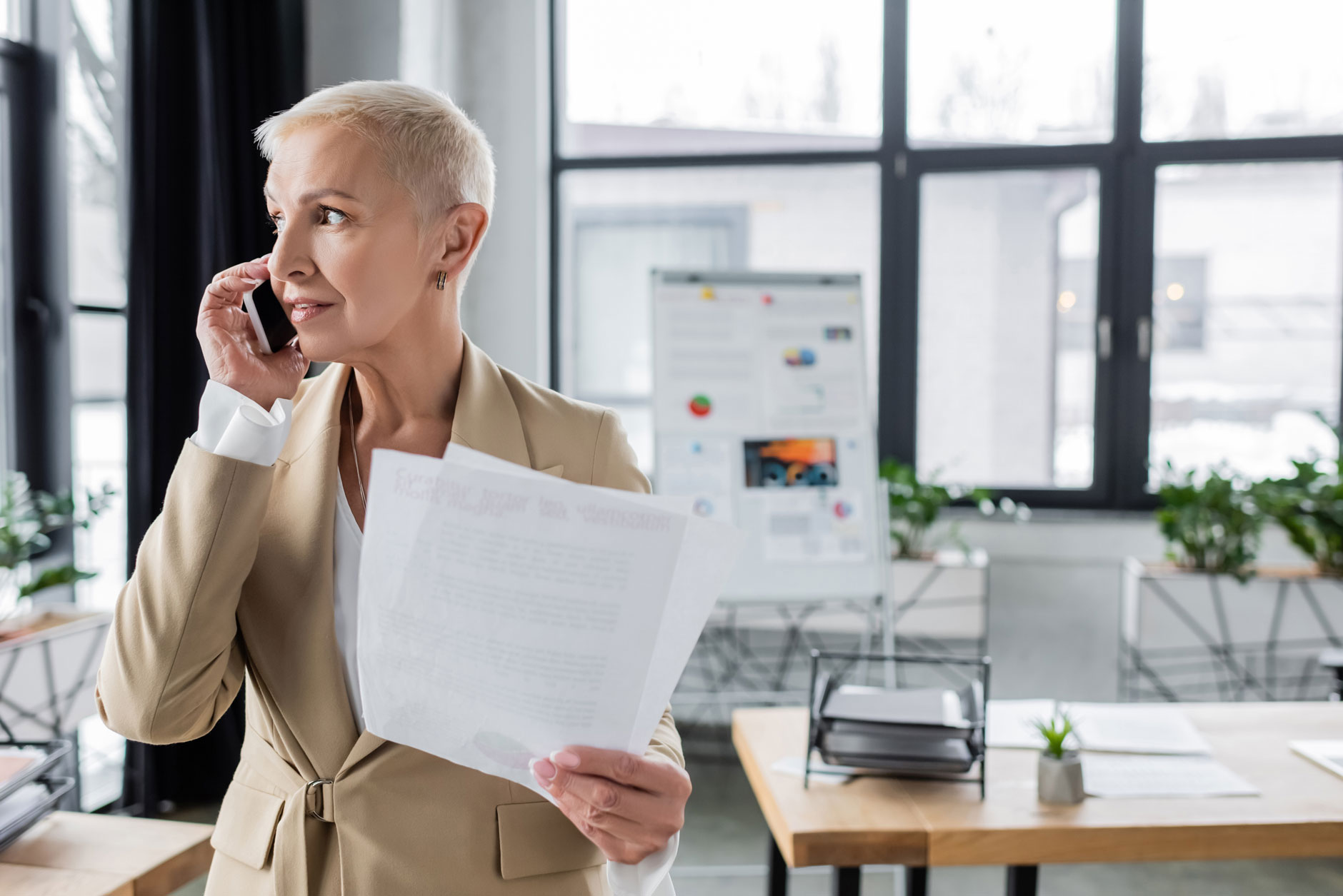 A woman with short blonde hair in a beige blazer holds documents and talks on her cellphone in a modern office with plants.