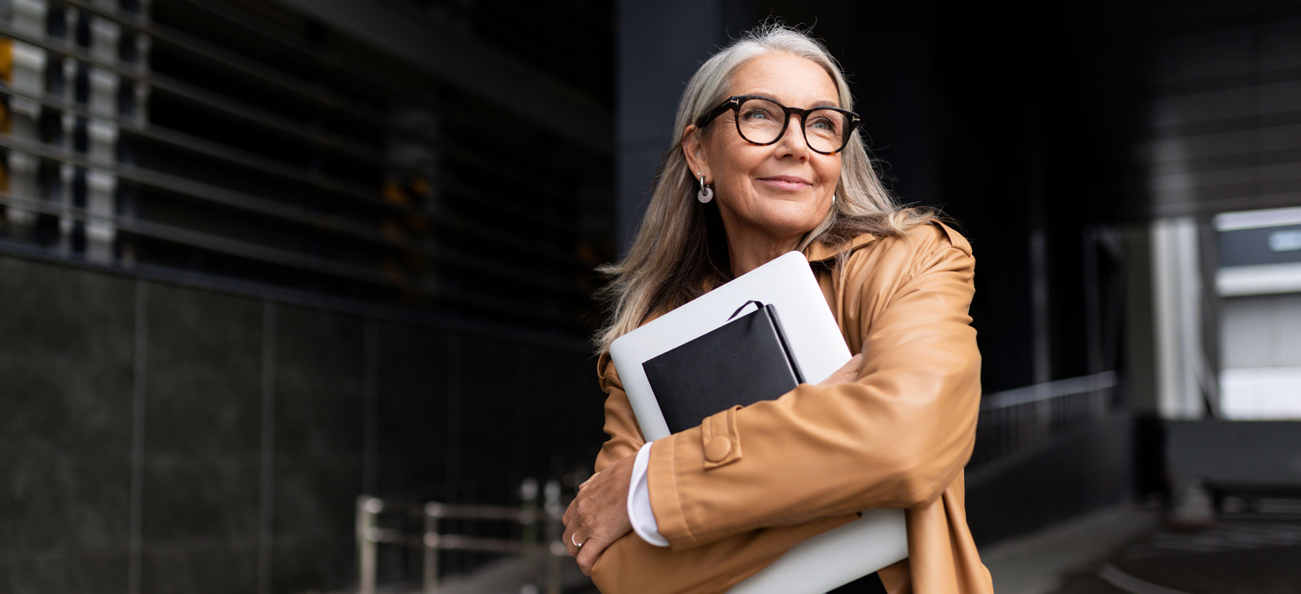 Older woman with gray hair and glasses in a tan coat stands outside, smiling, holding a closed laptop and black notebook.