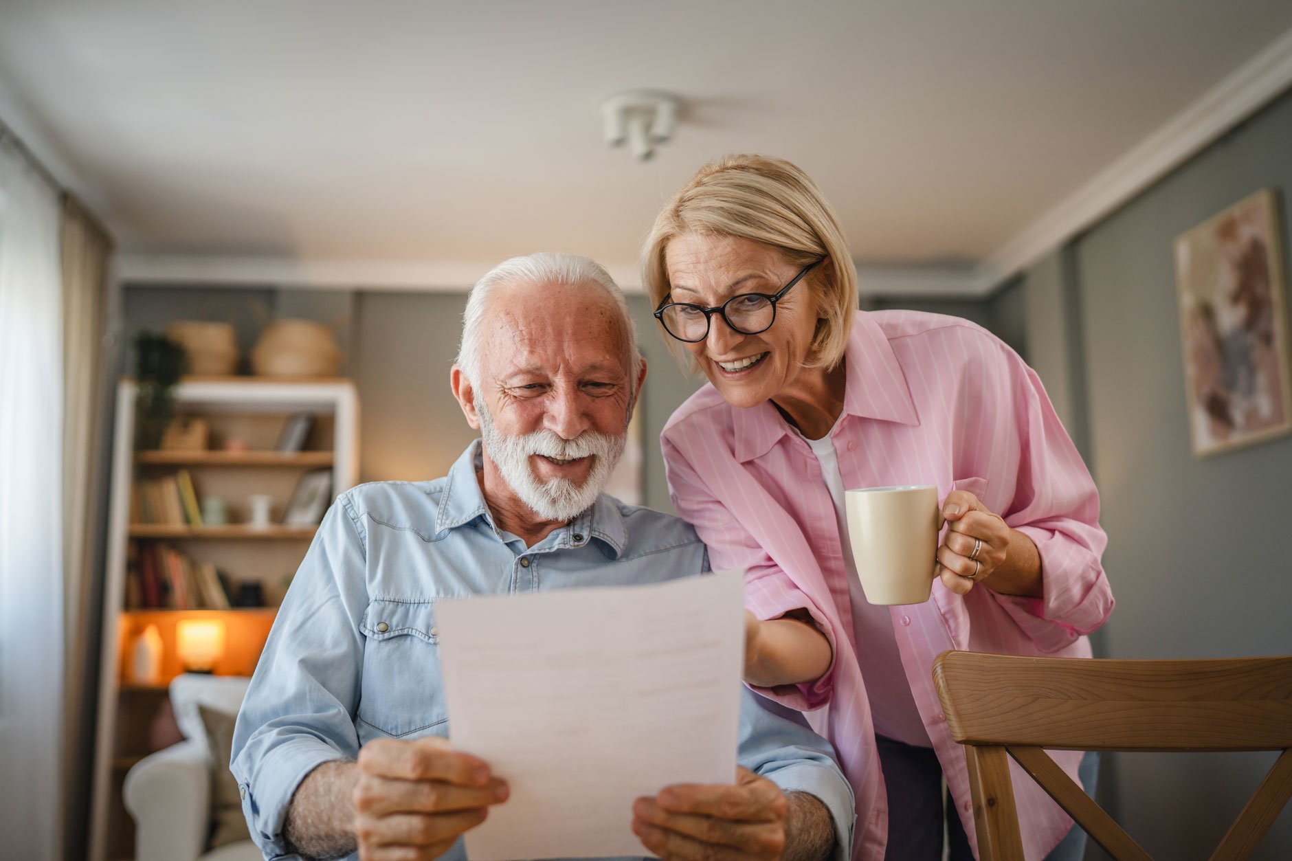 An elderly man and woman smile at a probate genealogy document in a cozy living room; he holds the paper, she holds a mug.