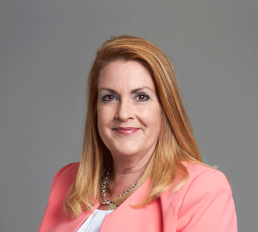 A woman with straight, shoulder-length reddish-blonde hair wears a coral blazer and smiles slightly against a plain gray background.