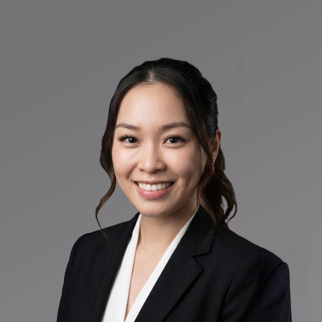 A woman with long dark hair in a ponytail, dressed in a black blazer and white blouse, smiles against a plain gray backdrop.