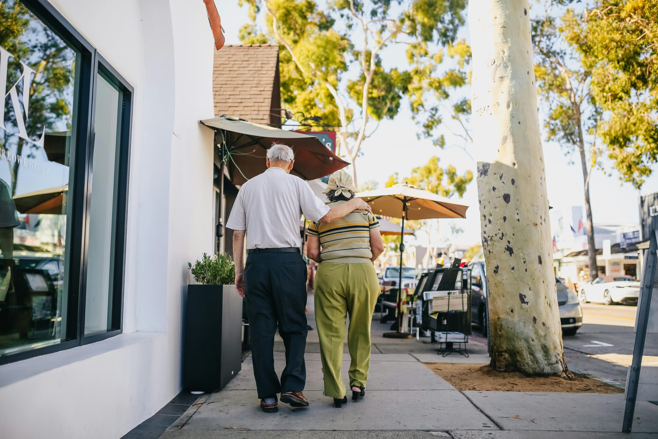 An elderly couple walks arm in arm along a sunny, tree-lined sidewalk beside outdoor cafes; the man holds the woman's shoulder.