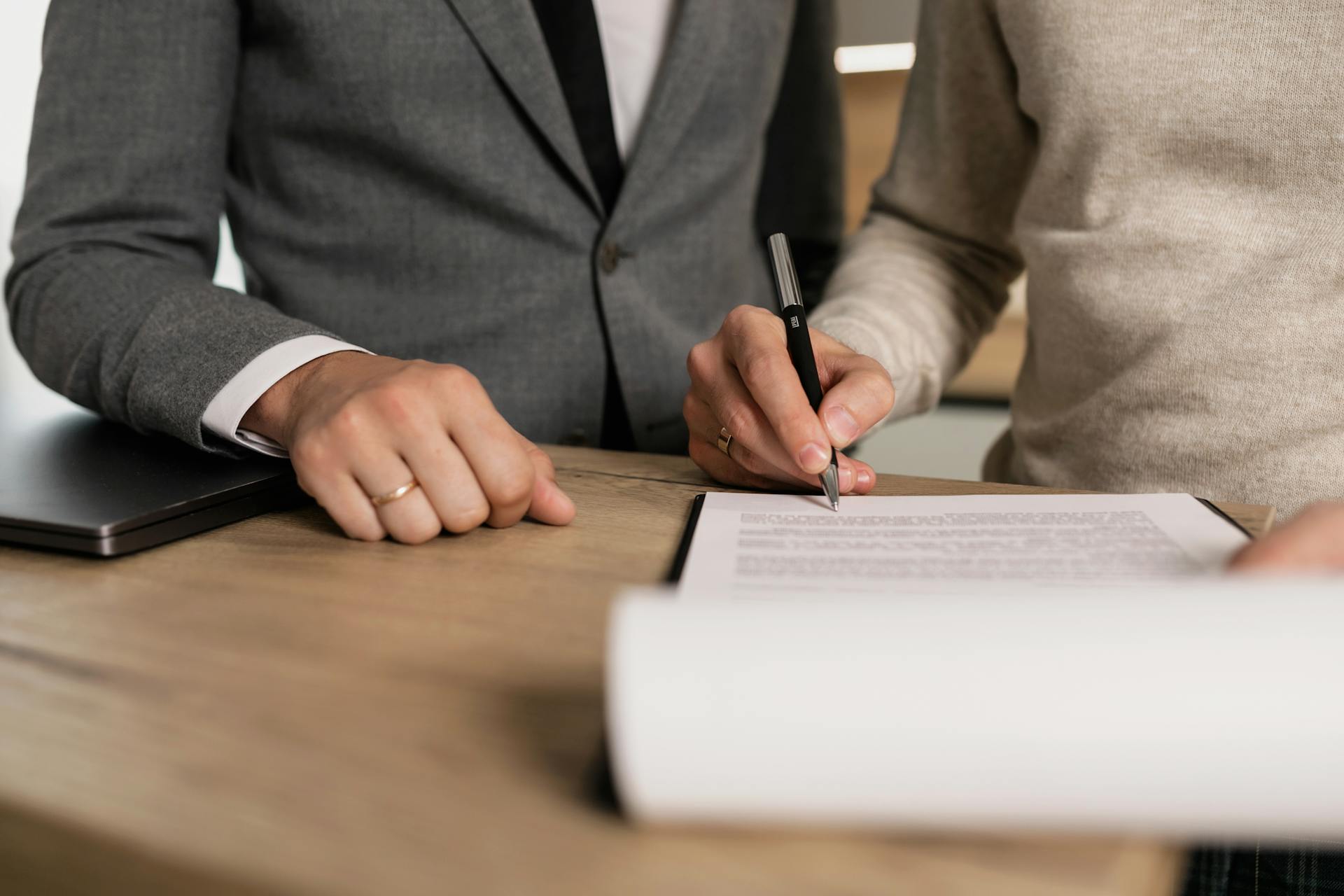 At a desk, a person in a beige sweater signs a document on a clipboard as someone in a gray suit watches.