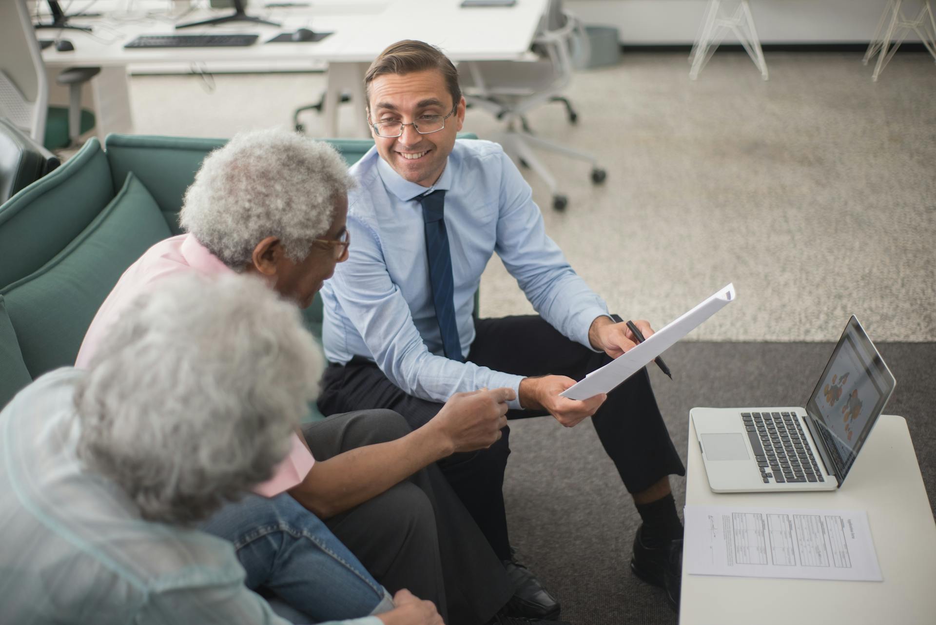 A man in a suit smiles and shows papers to two older adults sitting on a sofa, with a laptop on the coffee table nearby.
